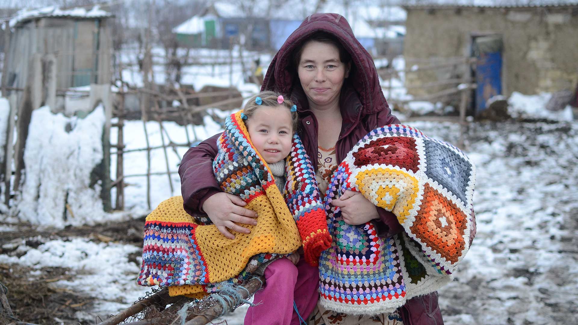 A woman stands with her child in the snow, with warm blankets.