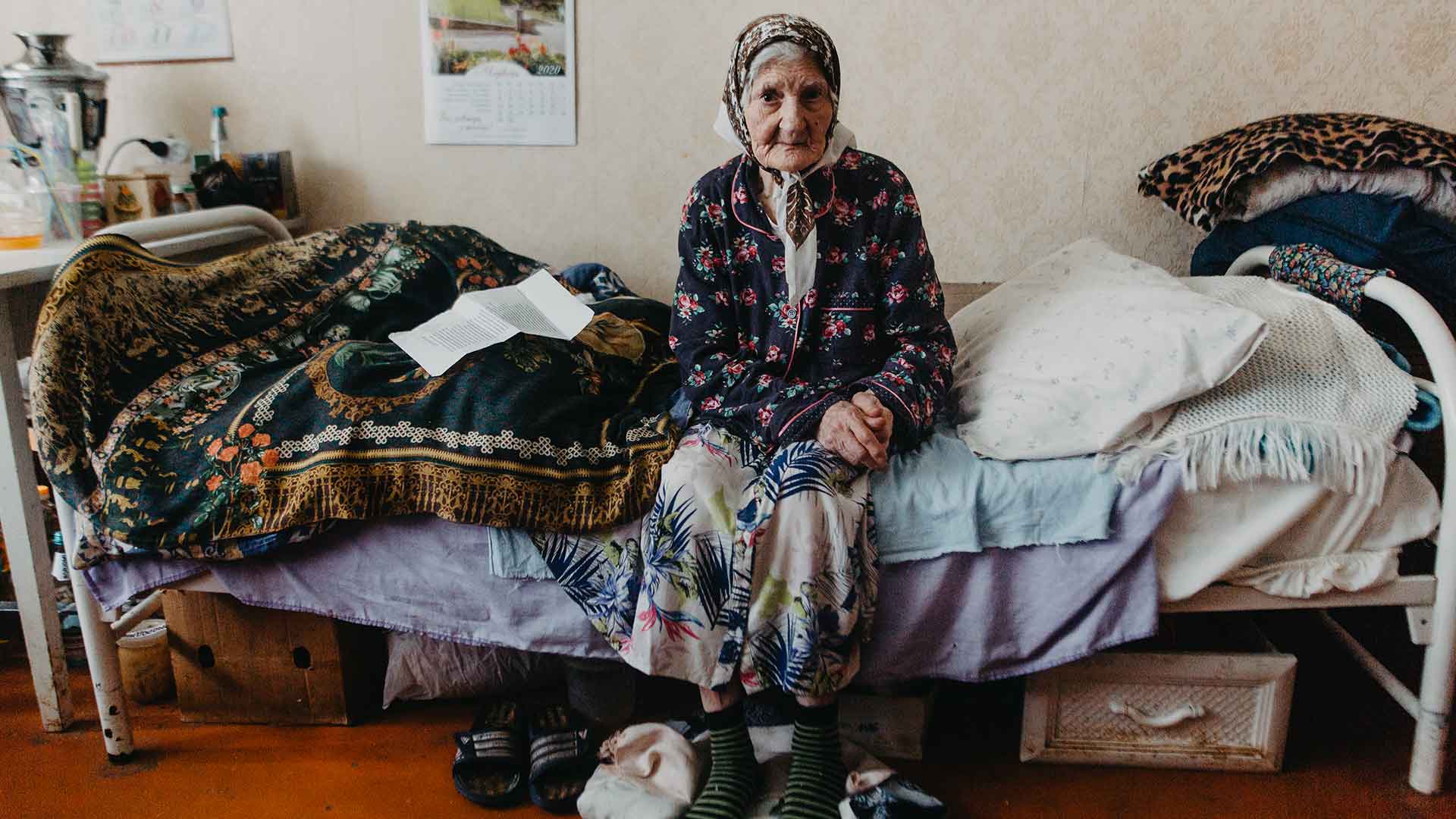 Elderly woman wearing a headscarf, sitting on a bed.