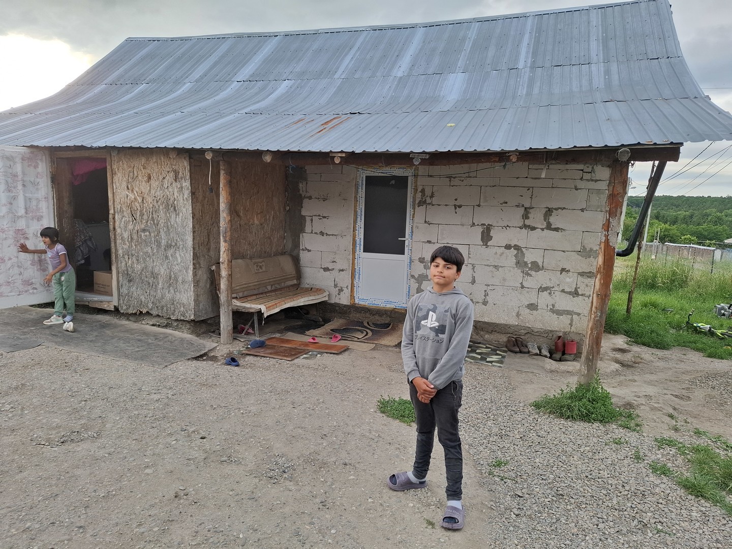 A boy standing in front of his small house with another child at the doorway.
