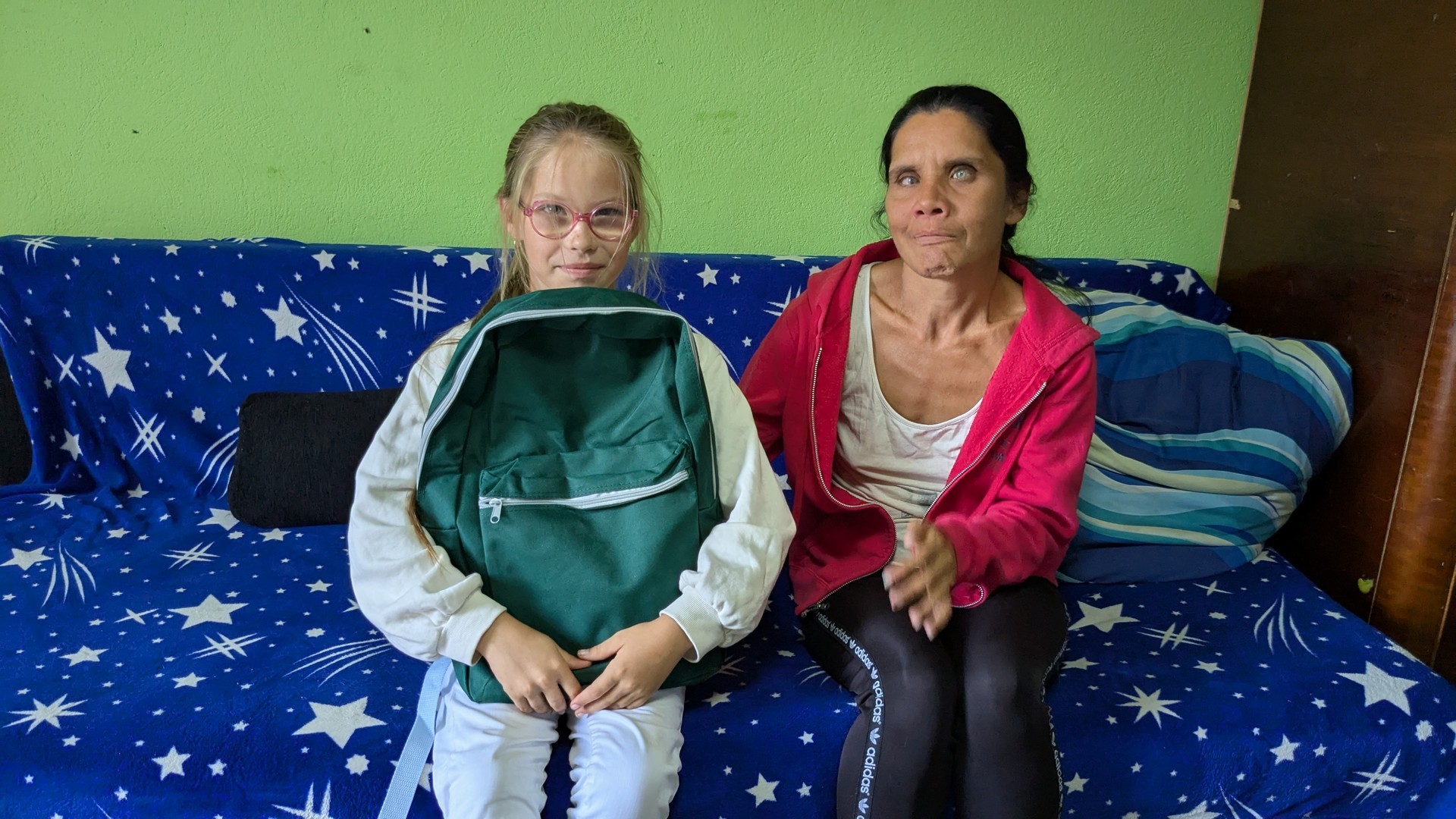 A girl holding her new backpack sitting on the couch with her mother.