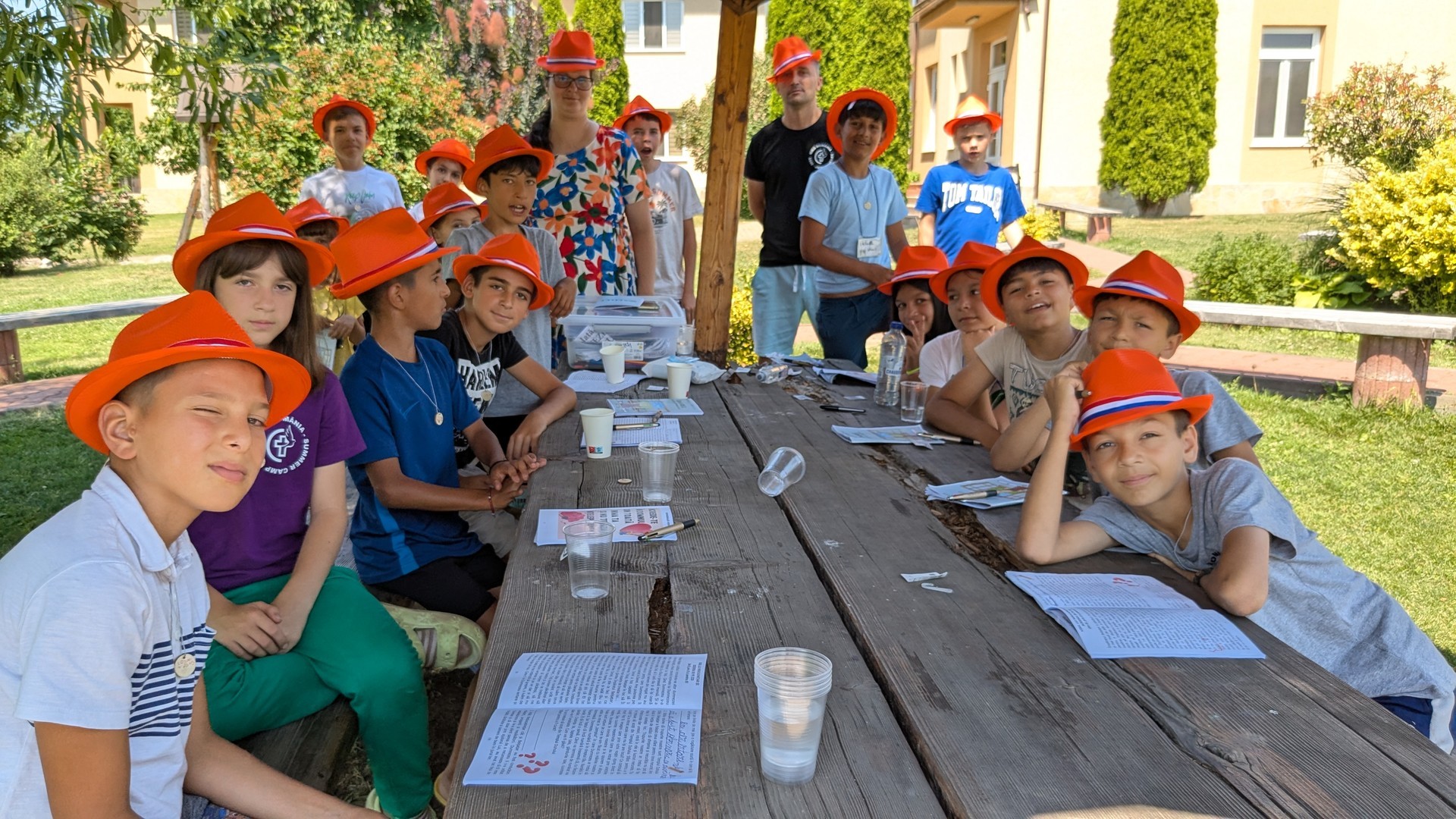 A bunch of kids and two adults sitting during summer camp at a picnic table with orange hats on